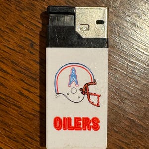 May include: A vintage lighter with a white front featuring a football helmet design in red, blue, and orange, with the word "OILERS" in red block letters. The top of the lighter is black and silver.