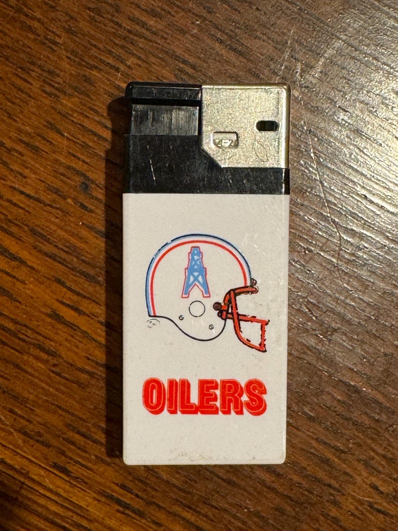 May include: A vintage lighter with a white front featuring a football helmet design in red, blue, and orange, with the word "OILERS" in red block letters. The top of the lighter is black and silver.