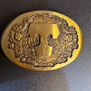 May include: An oval-shaped brass-toned belt buckle featuring a prominent capital letter T in the center. The background of the buckle is decorated with an intricate, raised floral and scrollwork pattern that surrounds the central letter.