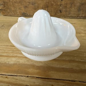 Könnte beinhalten: Weißer Glas-Zitruspresser mit konischem Reibkopf und einer Schüssel mit Ausguss. Der Entsafter besteht aus undurchsichtigem Glas und steht auf einer Holzoberfläche.