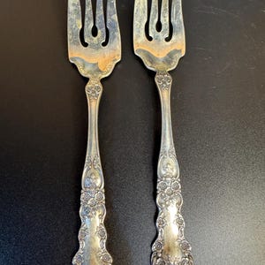 May include: Two silver-toned metal forks featuring ornate floral embossing along the handles and stems. The forks have three tines each, with a wide, flat base at the top of the handle where it meets the tines, showing signs of natural metal oxidation.