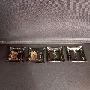 May include: Four square black glass ashtrays featuring gold-coloured graphics. Two ashtrays display a hotel building with the text HOTEL CASINO, while the other two depict a standing figure with the text LUCKY CASINO printed along the sides.