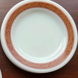 May include: White plate with a decorative orange and gold floral border. The plate has a simple, elegant design, suitable for serving food. The plate is round and appears to be made of ceramic.