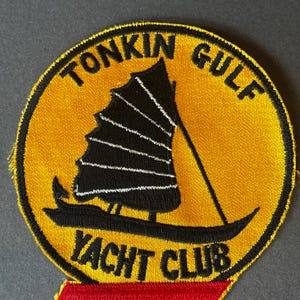 Könnte beinhalten: Gestickter Aufnäher mit gelbem Hintergrund und schwarzem Rand. Der Aufnäher zeigt ein schwarzes Segelboot mit weißem Segel und die Worte "TONKIN GULF YACHT CLUB" in schwarzer Schrift. Unten befindet sich ein roter rechteckiger Akzent.