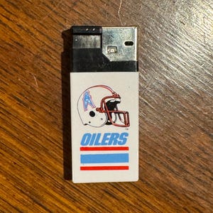 May include: A rectangular, white lighter with a black top. The lighter features a football helmet graphic with the word "OILERS" in blue text, and blue and red stripes. The lighter is on a wooden surface.