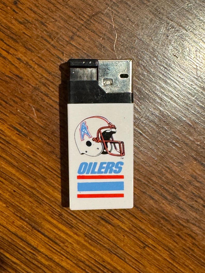May include: A rectangular, white lighter with a black top. The lighter features a football helmet graphic with the word "OILERS" in blue text, and blue and red stripes. The lighter is on a wooden surface.