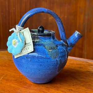 Könnte beinhalten: Handgefertigte blaue Keramik-Teekanne mit gebogenem Griff und Ausguss. Die Teekanne hat einen dunkelblauen Deckel und ein Etikett mit der Aufschrift "Jon's Pottery Barbados". Ein blaues blütenförmiges Etikett ist am Griff befestigt.
