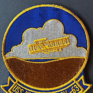 May include: Embroidered patch featuring a biplane flying over a brown and gray landscape, set against a blue sky and white clouds. The patch is bordered with a yellow trim and includes the text "USS KITTY HAWK CV-63" in yellow.
