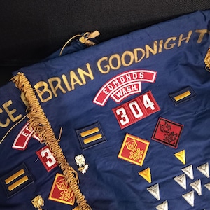 May include: A dark blue fabric banner featuring gold embroidered text reading BRIAN GOODNIGHT. It is decorated with various red, white, and blue Cub Scouts BSA patches, including the number 304, and several silver and gold metallic arrow-shaped pins.