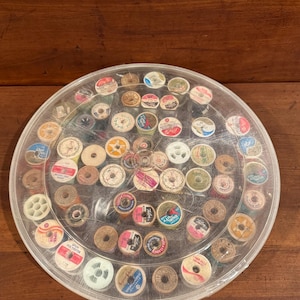 May include: A circular, clear plastic storage container filled with numerous small spools of sewing thread in various colors. The spools are arranged in concentric circles within the container, which features a transparent lid that allows for a clear view of the assorted thread brands and shades stored inside.