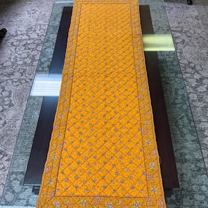 Könnte beinhalten: Rechteckiger, goldgelber, gesteppter Tischläufer mit Rautenmuster. Die Ränder sind mit einem floralen Bordürenmuster in Rot, Blau und Grün verziert. Der Läufer liegt auf einem dunkelbraunen Tisch mit Glasplatte.