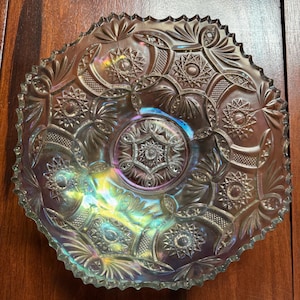 Puede incluir: Cuenco de vidrio iridiscente con un patrón detallado de estrellas, flores y formas geométricas. El cuenco tiene un borde festoneado y un diseño central en forma de estrella. El vidrio refleja un arcoíris de colores.