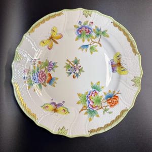 May include: White porcelain plate with a scalloped edge, decorated with colorful floral and butterfly designs. The plate features a green and gold trim, with a textured surface. A decorative piece for serving or display.