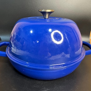 Puede incluir: Una olla redonda de hierro fundido esmaltado en color azul intenso con una tapa abovedada a juego. La olla cuenta con dos asas laterales y un pomo metálico central en la tapa. El exterior tiene un acabado brillante, liso y redondeado.