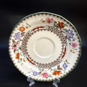 May include: A vintage saucer with a floral design. The white ceramic saucer features a brown, patterned border and is decorated with colorful flowers in shades of pink, orange, and blue. The rim is edged with green.