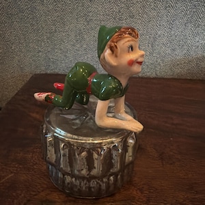 Op de afbeelding: Een vintage keramieken beeldje van een jongen in een groene outfit, knielend op een zilverkleurige container. De jongen heeft rood haar, roze wangen en kijkt naar rechts. De container heeft een decoratief, getextureerd ontwerp.