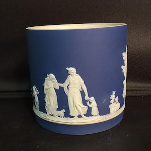 May include: A cylindrical blue ceramic vessel featuring white relief carvings of classical figures, including adults and children, arranged in a continuous scene around the exterior. The base and rim are finished with a thin, plain white border.