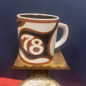 May include: A ceramic coffee mug with a white background and a brown and black abstract design. The number "78" is prominently displayed in the center. The mug sits on a vintage scale.