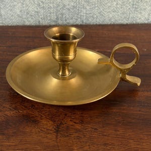 Könnte beinhalten: Ein Vintage-Kerzenhalter aus Messing mit rundem Sockel und Griff. Die Kerzenschale befindet sich mittig auf dem Sockel. Der Griff ist eine Schlaufe mit einer kleinen Lasche. Das Messing hat einen warmen, goldenen Farbton.