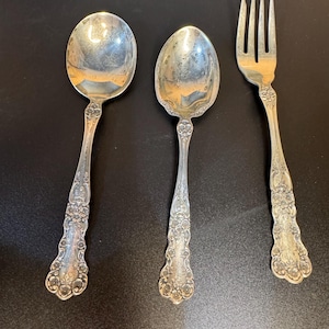 May include: Two silver-toned spoons and one silver-toned fork with ornate floral embossing on the handles. The utensils feature a polished finish with intricate decorative patterns extending from the base of the bowls and tines down to the handle ends.