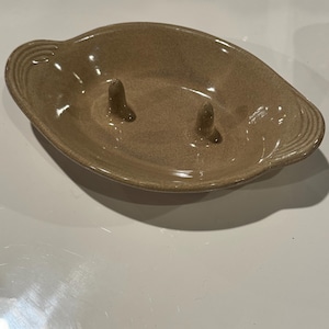 May include: A tan ceramic ashtray with a unique design. The ashtray is oval-shaped with two raised posts in the center and a ridged edge. The neutral color and simple design make it a versatile piece for any space.