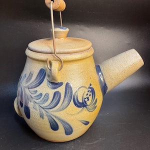 May include: A tan ceramic teapot with a lid and spout. The teapot features blue floral designs and a metal handle with a wooden grip. The teapot is likely vintage or handmade.