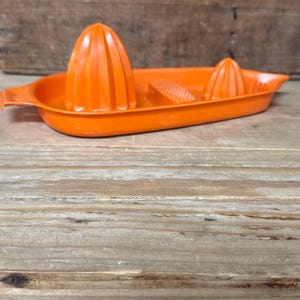 Op de afbeelding: Een oranje plastic citruspers met een ruimer en een kleine tuit. De citruspers heeft een grote, kegelvormige ruimer en een kleinere. De citruspers staat op een dienblad met een handvat.