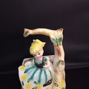 May include: A ceramic figurine planter featuring a blonde character with a teal and white dress adorned with yellow flowers, seated beneath a beige tree branch with a green butterfly and small red berries, all finished in a glossy glaze.