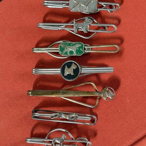 May include: A collection of seven silver and gold-toned metal tie clips, each featuring a distinct dog motif. The designs include various breeds in standing or running poses, some set against green or black enamel backgrounds or within circular frames.