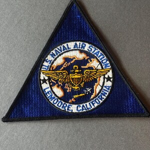 Könnte beinhalten: Ein dreieckiger Aufnäher mit marineblauem Hintergrund und schwarzem Rand. Der Aufnäher zeigt ein kreisförmiges Emblem mit dem Text "U.S. NAVAL AIR STATION LEMOORE, CALIFORNIA" um ein gold-weißes Design aus Flügeln und einem Globus.