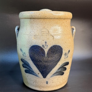 May include: A tan stoneware crock with a lid, featuring a large blue heart design. The crock has two small handles and the text "ROWAN POTTERY" is visible. A classic piece of kitchenware.