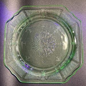 May include: A vintage green glass plate with an octagonal shape and a decorative floral and scroll pattern. The plate has a raised rim and a central design, perfect for serving appetizers or desserts.
