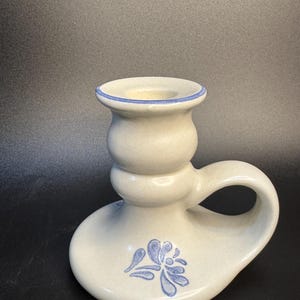 Könnte beinhalten: Cremefarbener Kerzenhalter aus Keramik mit blauem Blumenmuster. Der Kerzenhalter hat einen abgerundeten Sockel, einen Griff und eine blau umrandete Öffnung für eine Kerze. Das Design ist klassisch und rustikal.