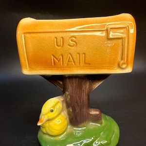 May include: A ceramic mailbox-shaped figurine with the words "US MAIL" in brown lettering. The mailbox is supported by a brown tree trunk and a green base with a yellow duckling.