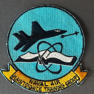 Könnte beinhalten: Runder, gestickter Aufnäher mit türkisfarbenem Hintergrund. Er zeigt ein schwarzes Flugzeug, ein weißes und schwarzes stilisiertes Atom und den Text "NAVAL AIR MAINTENANCE TRAINING GROUP" in Gelb. Der Aufnäher hat einen gelben Rand.