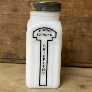 May include: Vintage white glass spice jar with a metal lid. The jar has the words "Purified Paprika" and "Griffiths'" printed in black. The jar is rectangular with vertical ridges.
