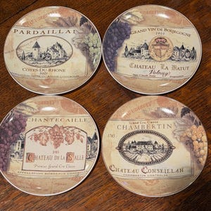 May include: Four ceramic coasters with wine label designs. Each coaster features a different vintage wine label with illustrations of chateaus, grapes, and text. The coasters have a cream-colored background and a brown border.