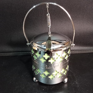 Può includere: Un barattolo per conserve in metallo cromato con manico oscillante, coperchio con fessura per cucchiaio e un motivo decorativo traforato che rivela un inserto in vetro verde pallido. La parola Preserves è impressa sul davanti del rivestimento.