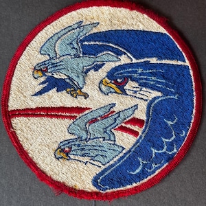Könnte beinhalten: Runder, gestickter Aufnäher mit rotem Rand, der drei stilisierte Adler zeigt. Die Adler sind hellblau mit roten und gelben Details, vor einem cremefarbenen Hintergrund. Das Design deutet auf ein militärisches oder luftfahrtbezogenes Thema hin.