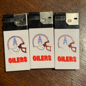 May include: Three white lighters with a football helmet design in red, blue, and white. The word "OILERS" is printed in red below the helmet graphic. The lighters have a silver top.