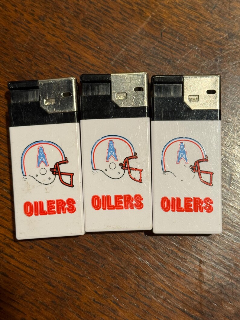 May include: Three white lighters with a football helmet design in red, blue, and white. The word "OILERS" is printed in red below the helmet graphic. The lighters have a silver top.