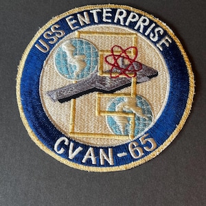 Könnte beinhalten: Gestickter Aufnäher mit dem Text "USS ENTERPRISE CVAN-65". Der Aufnäher hat einen blauen Rand mit goldenen Zierleisten, der ein beiges Zentrum mit einem grau-roten Design und zwei blau-weißen Globen umgibt.