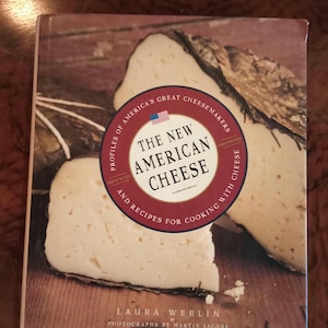 以下が含まれることがあります： ローラ・ワーリン著『The New American Cheese: Profiles of America's Great Cheesemakers and Recipes for Cooking with Cheese』というタイトルのハードカバー本。表紙には2切れのチーズの写真と、小さなアメリカ国旗のアイコンが描かれています。