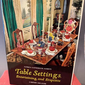May include: A book titled "Table Settings, Entertaining, and Etiquette" by Patricia Easterbrook Roberts. The cover features a dining table set with red glassware, candles, and grapes. The background shows a room with green curtains and ornate chairs.