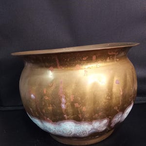 May include: A rounded brass planter with a wide, flared rim. The metal surface displays a warm golden tone with visible vertical streaks of oxidation, including patches of green patina near the base and scattered copper-colored spots throughout.