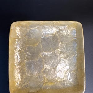 May include: A square decorative tray with rounded corners. The tray is covered in iridescent, overlapping, circular pieces, creating a shimmering, textured surface. The tray has a light, golden-yellow hue, with a dark background.