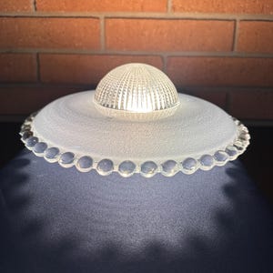 Może przedstawiać: Przezroczysty szklany klosz lampy z matową, w kształcie spodka podstawą i małą, kopulastą górą. Podstawa ma falisty brzeg. Góra to mała, żebrowana kopuła. Klosz lampy jest podświetlany od wewnątrz, tworząc delikatną poświatę.