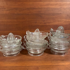 Peut inclure: Une collection de presse-agrumes en verre transparent disposés en trois piles. Chaque presse-agrumes présente un cône central, un bol nervuré pour recueillir le jus et une petite poignée latérale. Le verre est transparent avec des stries verticales.