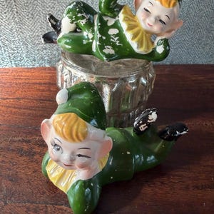 May include: Two vintage ceramic elf figurines, one winking, both in green outfits with yellow collars and hats. The elves are lying down on a clear glass container, with black shoes and white accents. The figurines have a whimsical, antique aesthetic.