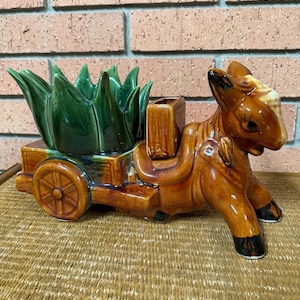 May include: A glazed ceramic planter shaped like a brown donkey pulling a cart. The cart features a dark green, spiky, leaf-like container. The donkey has black-painted hooves, a white patch on its forehead, and a small rectangular saddle pack.